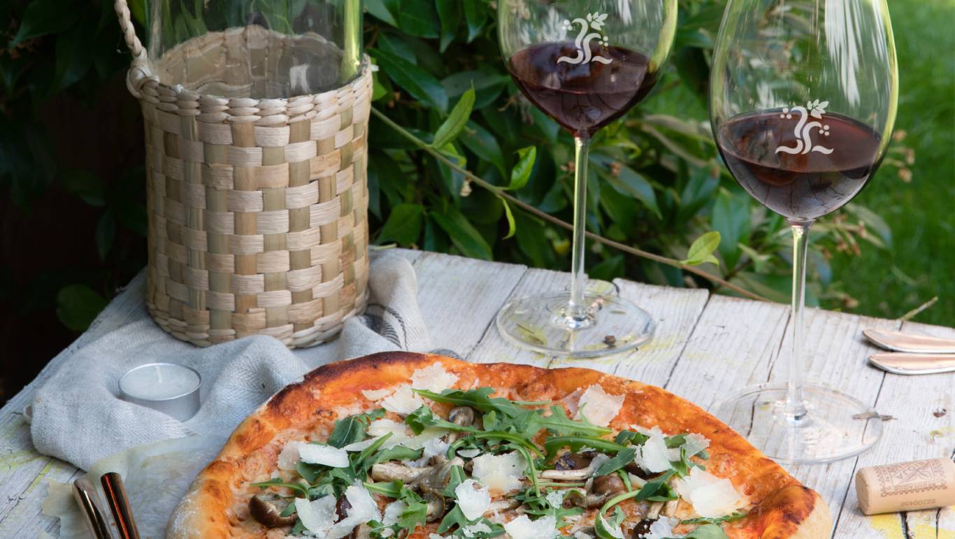 Two glasses of red wine on a table outdoors by a pizza
